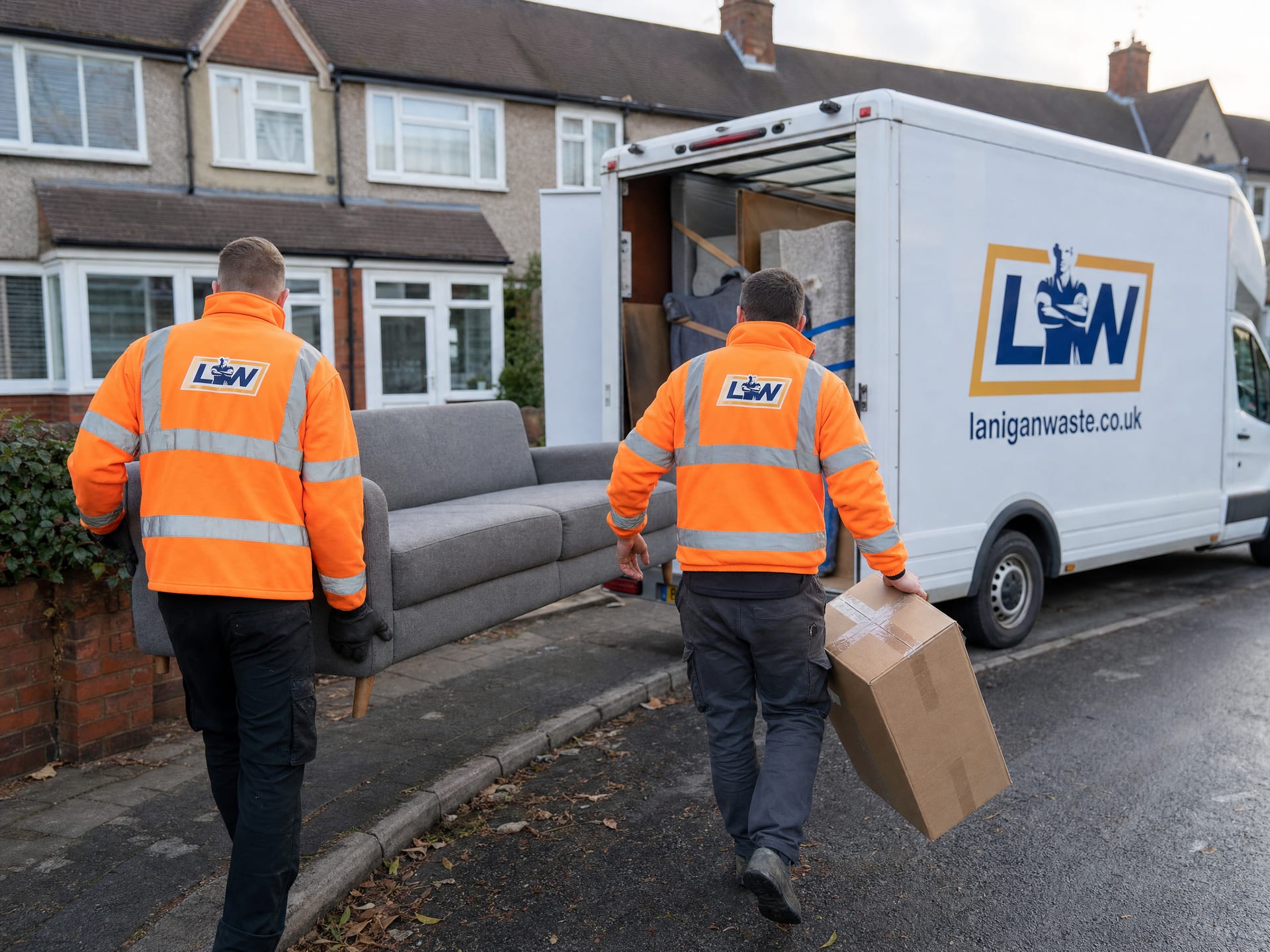 Two Lanigan Waste crew in hi-vis carrying a sofa and a box to the branded van outside a house